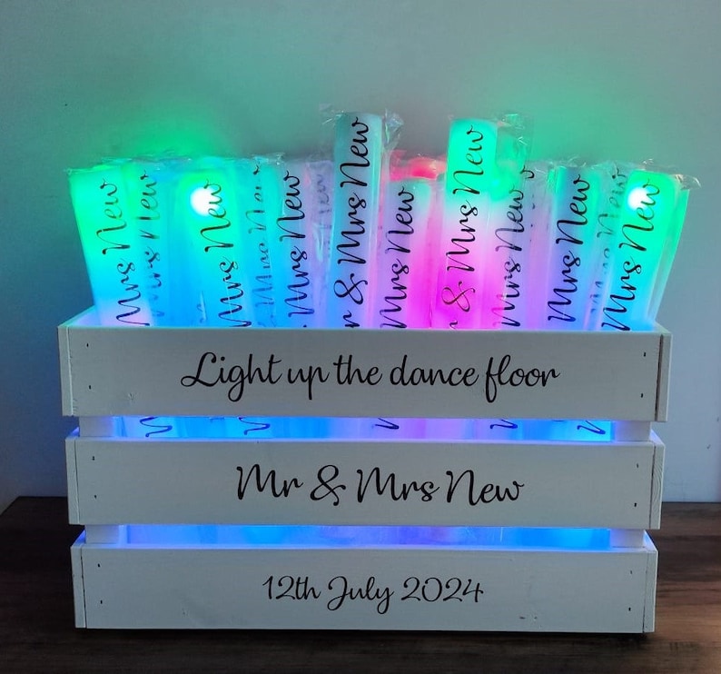 May include: A white wooden crate with multi-colored LED lights, holding glowing light sticks. The crate has the text "Light up the dance floor", "Mr & Mrs New", and "12th July 2024". The light sticks are labeled "Mrs New".