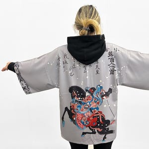 May include: Gray Japanese-style kimono with a black hood, featuring a colorful illustration of a warrior riding a horse. The kimono has Japanese characters printed on the back.