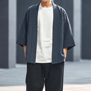 May include: A navy blue kimono-style jacket with wide sleeves, worn over a white t-shirt and paired with black trousers. The jacket has a relaxed fit and a simple, open front design. The outfit is casual and modern.