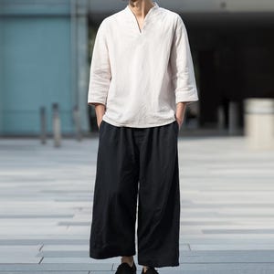 May include: A person wearing a cream-colored, V-neck shirt with three-quarter sleeves and black wide-leg pants. The outfit is a casual, minimalist style. The person is wearing black shoes.