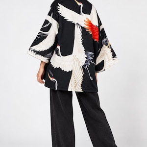 Könnte beinhalten: Eine schwarze Jacke im Kimono-Stil mit einem wiederkehrenden Druck von weißen Kranichen mit roten Kämmen und orangefarbenen Flügelspitzen, kombiniert mit einer locker sitzenden schwarzen Jeanshose und weißen Turnschuhen. Die Jacke hat weite Ärmel.