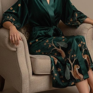 May include: A dark green velvet robe with a tie waist and wide sleeves. The robe features an embroidered design of trees and tigers in shades of gold and brown. The robe is worn while seated in a beige armchair.