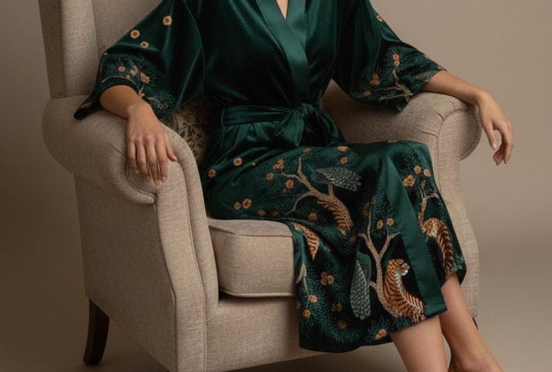 May include: A dark green velvet robe with a tie waist and wide sleeves. The robe features an embroidered design of trees and tigers in shades of gold and brown. The robe is worn while seated in a beige armchair.