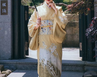 May include: A woman wearing a pale yellow kimono with floral patterns and a decorative obi. The kimono features white floral designs and a contrasting patterned obi. The sleeves are long and flowing. The woman is holding a black umbrella.
