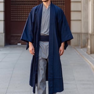 Puede incluir: Una bata azul marino estilo kimono con mangas anchas, usada sobre un atuendo gris. La bata es larga, llega hasta los tobillos, y se asegura con una faja negra. El atuendo es una mezcla de estilos tradicionales y modernos.