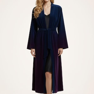 May include: A long, velvet robe in a gradient of blue to purple. The robe has a tie belt and wide sleeves. The model is wearing a black slip underneath. The robe is a luxurious piece of loungewear.