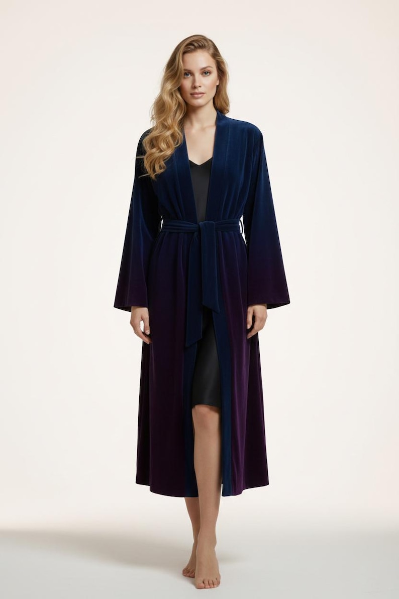 May include: A long, velvet robe in a gradient of blue to purple. The robe has a tie belt and wide sleeves. The model is wearing a black slip underneath. The robe is a luxurious piece of loungewear.