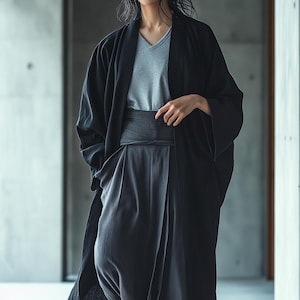 Puede incluir: Una bata negra estilo kimono con mangas anchas, usada sobre una camiseta gris con cuello en V y pantalones grises oscuros. Una faja ancha gris oscura ciñe la cintura. El atuendo es minimalista y moderno, con un enfoque en líneas fluidas y tejidos cómodos.