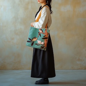 以下が含まれることがあります： 鳥と花の模様が特徴の、ターコイズ色のトートバッグ。バッグは長方形で、ライトブラウンのストラップと白と黒の服装です。アートワークは、白い花のある枝に止まっている2羽の鳥を描いています。