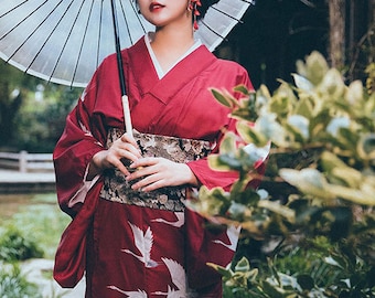 May include: A burgundy kimono with white crane designs and a decorative obi belt. The traditional Japanese garment is worn with a white parasol. The kimono's sleeves are long and flowing, and the obi belt is black with gold floral patterns.