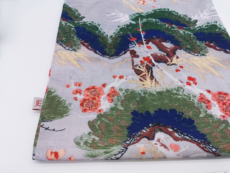 May include: A gray fabric tote bag with a floral print featuring red, white, and green flowers and leaves. The bag has a white background with a subtle texture. The bag has a small red label with the letters "JE" on it.