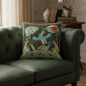Può includere: Cuscino quadrato decorativo con un design ricamato raffigurante un uccello blu, onde, nuvole e un sole rosso su sfondo scuro. Il cuscino è posto su un divano in pelle verde, aggiungendo un tocco di eleganza.