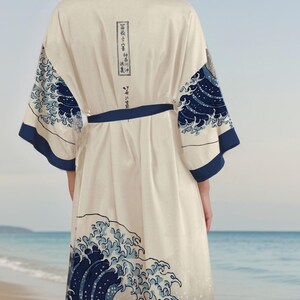 May include: A white robe with a blue and white wave pattern. The robe has a tie belt and long sleeves. The wave pattern is inspired by the famous Japanese woodblock print "The Great Wave off Kanagawa" by Katsushika Hokusai. The robe is shown against a beach background.