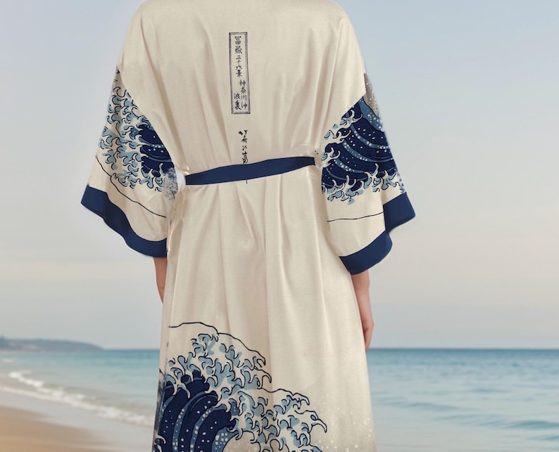 May include: A white robe with a blue and white wave pattern. The robe has a tie belt and long sleeves. The wave pattern is inspired by the famous Japanese woodblock print "The Great Wave off Kanagawa" by Katsushika Hokusai. The robe is shown against a beach background.