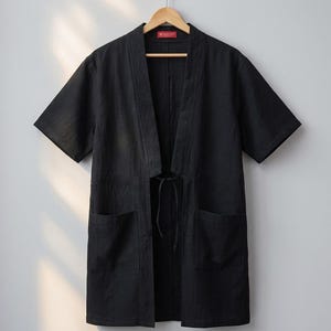 Może przedstawiać: Czarna, rozpinana z przodu kurtka w stylu kimono z kr&oacute;tkimi rękawami, szerokim kołnierzem i dwiema dużymi naszywanymi kieszeniami z przodu. Odzież posiada cienkie czarne wiązanie w talii i jest wyeksponowana na drewnianym wieszaku na ubrania.