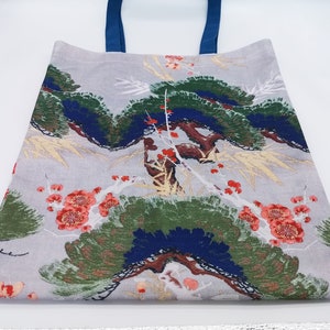 May include: A gray tote bag with blue handles and a floral print featuring red flowers, green leaves, and blue and gold accents. The bag has a small red label with the text "JE" on it.