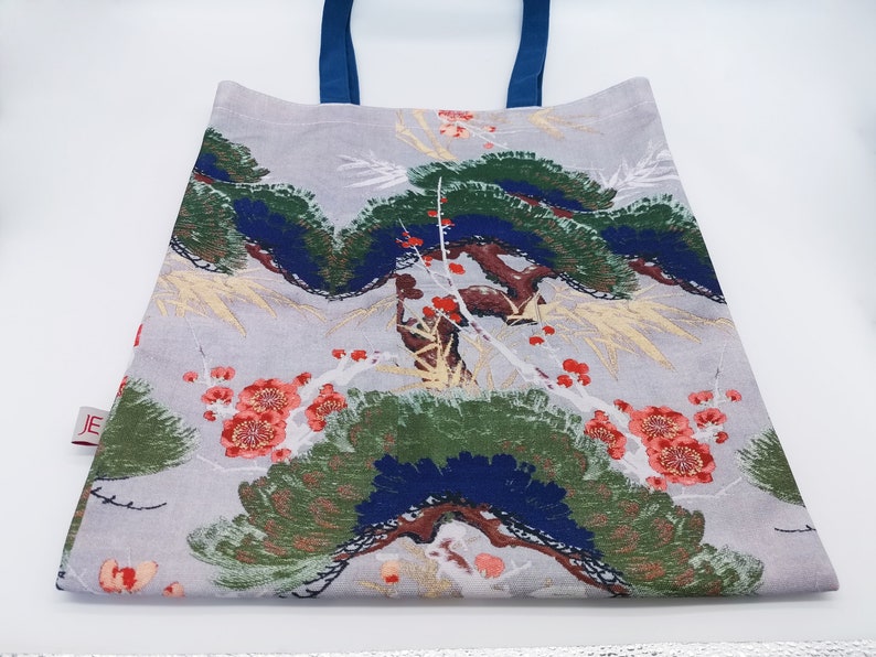 May include: A gray tote bag with blue handles and a floral print featuring red flowers, green leaves, and blue and gold accents. The bag has a small red label with the text "JE" on it.