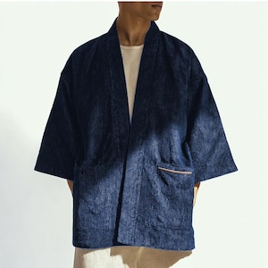 Peut inclure: Veste kimono en velours côtelé bleu marine avec une coupe décontractée. La veste présente un col châle, des manches trois-quarts et deux poches avant avec une bordure beige. Le modèle porte une chemise de couleur crème en dessous.