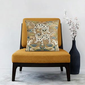 May include: A decorative throw pillow with an embroidered design of two spotted animals on a tan background. The pillow is placed on a mustard yellow chair with dark wood legs. A black vase with white flowers is to the right.