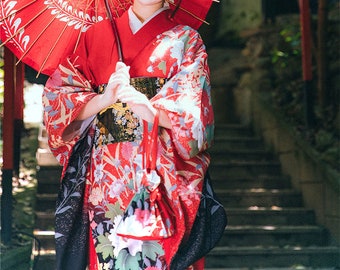 May include: A person wearing a vibrant red kimono with floral patterns, holding a matching red parasol and a small drawstring bag. The kimono features intricate designs in red, white, and black, creating a striking visual contrast. The overall aesthetic suggests traditional Japanese attire.