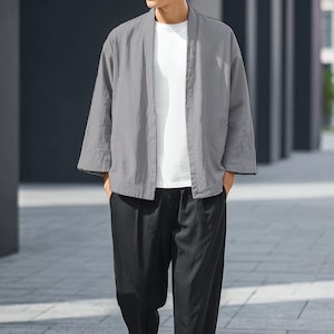 May include: A grey, open-front kimono-style jacket with three-quarter sleeves. The jacket is worn over a white t-shirt and paired with black trousers. The outfit is casual and appears to be made of a lightweight fabric.
