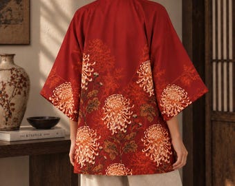 May include: A deep red kimono-style jacket featuring a dark brown collar and a vibrant floral pattern of orange and yellow chrysanthemums. The garment has wide, three-quarter length sleeves and is paired with loose-fitting, light beige trousers.
