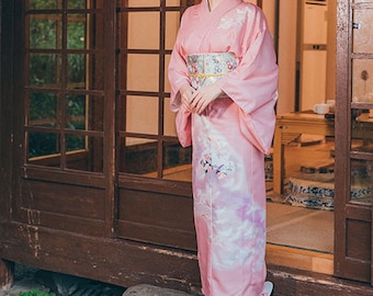 May include: A pink kimono with a floral design on the lower half and a patterned obi. The sleeves are long and flowing. The kimono is worn in front of a traditional Japanese wooden structure.