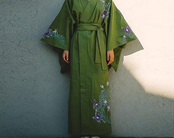 May include: Olive green kimono with embroidered purple flowers and butterflies. The kimono features wide sleeves, a wrap-around design, and a matching belt. The garment is worn with white sandals.