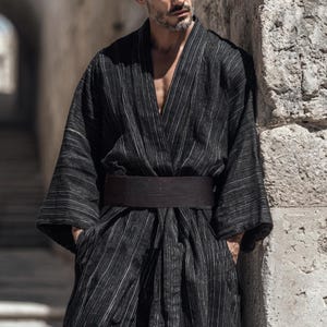 May include: A black kimono-style robe with vertical white stripes and a wide, dark brown belt. The robe has wide sleeves and a deep V-neck. The person is leaning against a stone wall.