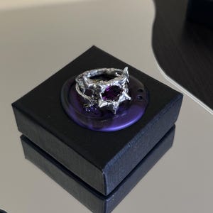 May include: A silver ring with a deep purple gemstone, presented on a dark purple wax seal. The ring sits on a black square box, reflecting on a mirrored surface. The ring's design features detailed, textured elements.