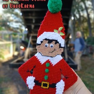 May include: A handmade, flat elf ornament with a red suit, white collar and cuffs, and a green pom-pom on the hat. The elf has a black belt with a gold buckle and green buttons. Text on the ornament reads "Flat Elf" and "An Original Design by UndoKarma."
