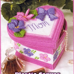 May include: A pink heart-shaped plastic canvas box with a lid, decorated with purple and pink flowers, a purple bow, and the word "Mom". The box has a white border with floral designs. The text "Plastic Canvas PDF Pattern Download" is at the bottom.