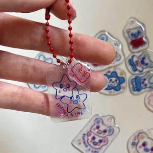 Könnte beinhalten: Ein klarer Acryl-Schlüsselanhänger mit zwei blauen Sternfiguren und dem Schriftzug "FRIENDS 4 LIFE" in Schwarz. Der Schlüsselanhänger ist an einer roten Kugelkette befestigt. Weitere kleine, klare Acryl-Anhänger mit Cartoon-Figuren befinden sich im Hintergrund.