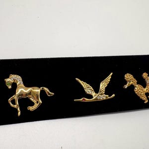 Op de afbeelding: Drie goudkleurige metalen broches met een paard met een groen stenen oog en heldere kristallen, een vliegende kraanvogel met heldere kristallen en een rood stenen oog, en een gestructureerde poedel met heldere kristallen op de kop en staart.