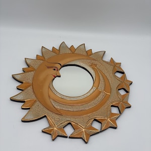 Op de afbeelding: Een ronde wandspiegel met een decoratieve lijst in de vorm van een maansikkel met een gezicht en omringende zonnestralen. De lijst heeft een textuur met een craquelé afwerking in beige en goudtinten met kleine sterren.