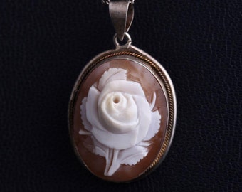 May include: Oval pendant necklace with a carved white rose cameo set in a silver-tone frame and chain. The cameo is set against a brown background, creating a contrast. The jewellery is displayed against a dark background.