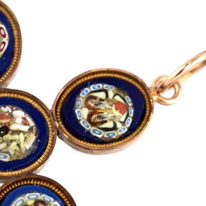 May include: A cross-shaped pendant crafted from gold-toned metal, featuring multiple oval settings. Each setting contains a deep blue background with intricate micro-mosaic glass floral patterns in white, red, and black, finished with a jump ring.