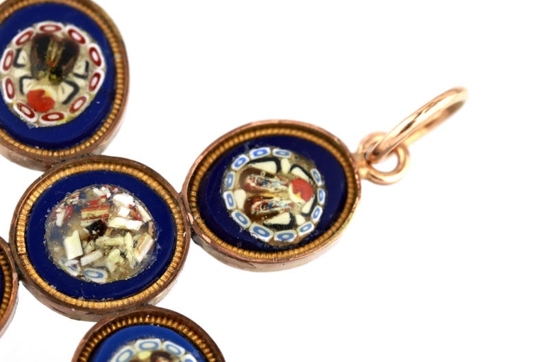 May include: A cross-shaped pendant crafted from gold-toned metal, featuring multiple oval settings. Each setting contains a deep blue background with intricate micro-mosaic glass floral patterns in white, red, and black, finished with a jump ring.