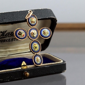 May include: A gold-coloured cross pendant with circular, detailed insets. Each inset features a central design surrounded by blue enamel and gold trim. The cross is displayed in a black box with a blue velvet lining.