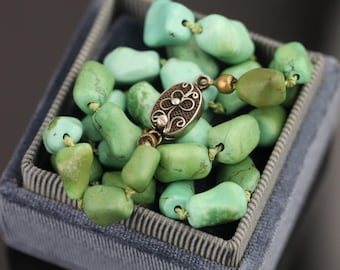 May include: A necklace of turquoise beads with a decorative silver clasp sits in a blue velvet box. The beads are various shades of green, and the box has a striped border.