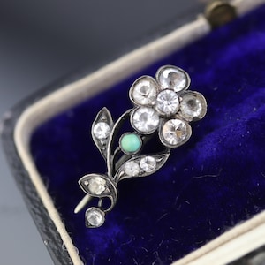 May include: A silver-toned metal brooch shaped like a flower with a stem and leaves. The flower petals and leaves are set with clear, faceted rhinestones. A single, small, round, opaque turquoise-colored stone is positioned on the stem.