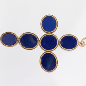 May include: A gold-coloured pendant in the shape of a cross, with oval, deep blue gemstones. The gemstones are set within a gold frame. The pendant has a small loop at the top for attaching to a chain.