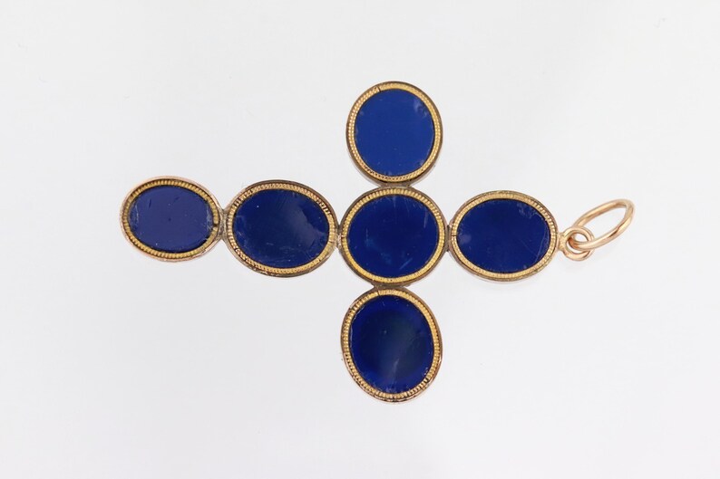 May include: A gold-coloured pendant in the shape of a cross, with oval, deep blue gemstones. The gemstones are set within a gold frame. The pendant has a small loop at the top for attaching to a chain.