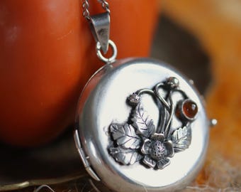 May include: A round silver locket pendant on a delicate chain, featuring a raised floral motif with textured leaves and a small, round, reddish-brown gemstone set into the metal surface. The locket has a polished finish and a hinged closure.