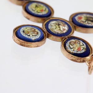 May include: A cross-shaped pendant featuring five circular micro-mosaic insets. Each inset displays a floral pattern in white, yellow, and red against a deep blue background, all encased in a metallic gold-toned frame with a small bail.