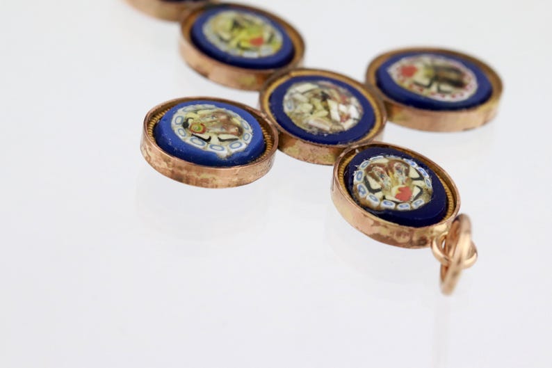 May include: A cross-shaped pendant featuring five circular micro-mosaic insets. Each inset displays a floral pattern in white, yellow, and red against a deep blue background, all encased in a metallic gold-toned frame with a small bail.