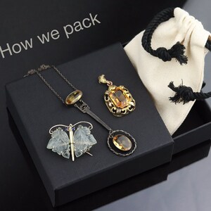 May include: A black box with a white fabric bag inside. The box contains three pieces of jewellery: a butterfly brooch, a pendant necklace, and a pendant on a chain. The text "How we pack" is written on the top of the box.