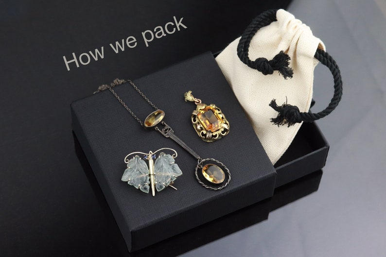 May include: A black box with a white fabric bag inside. The box contains three pieces of jewellery: a butterfly brooch, a pendant necklace, and a pendant on a chain. The text "How we pack" is written on the top of the box.