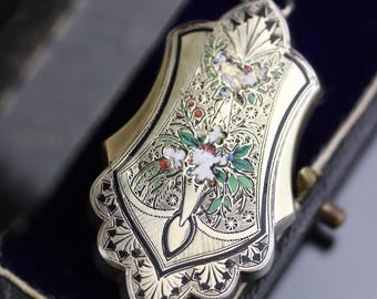 May include: A gold-toned metal pendant with an ornate, shield-like shape featuring intricate black engraved detailing and colourful floral enamel accents in red, green, and white. The piece includes a small circular bail at the top for hanging.
