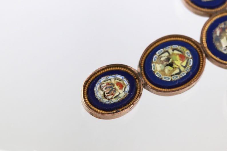 May include: A bracelet composed of oval links featuring deep blue enamel borders and central micro-mosaic floral patterns. Each link is framed in a metallic setting with a beaded edge, connected by small metal rings in a repeating sequence.